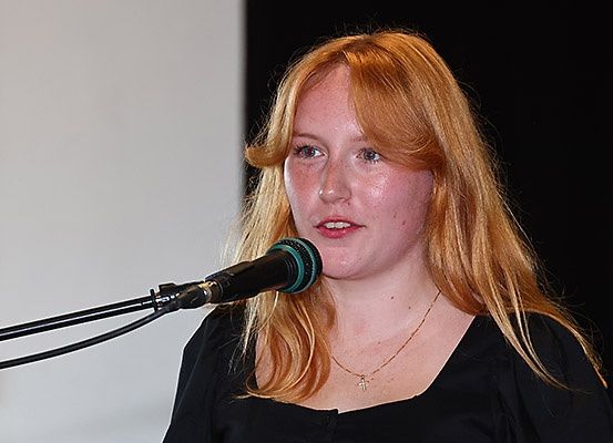 Eine Frau mit blonden Haaren spricht in ein Mikrofon. Sie trägt ein schwarzes Oberteil und eine Halskette. Das Mikrofon ist schwarz mit einer grünen Spitze. Sie steht vor einer weißen Wand.