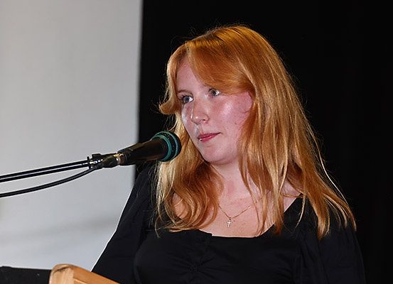 Eine Frau mit langen, welligen blonden Haaren spricht in ein Mikrofon an einem Podium. Sie trägt ein schwarzes Oberteil und eine Halskette.