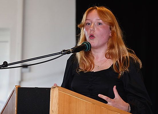 Eine Frau mit langen blonden Haaren steht an einem Rednerpult und spricht in ein Mikrofon. Sie trägt ein schwarzes Oberteil.