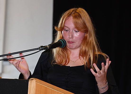 Eine Frau mit blonden Haaren steht an einem Rednerpult und spricht in ein Mikrofon. Sie trägt ein schwarzes Oberteil und hat eine Halskette und Armbänder an.