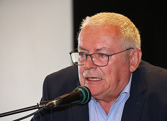 Ein älterer Mann mit kurzen blonden Haaren spricht in ein Mikrofon. Er trägt eine Brille und eine dunkle Anzugjacke.