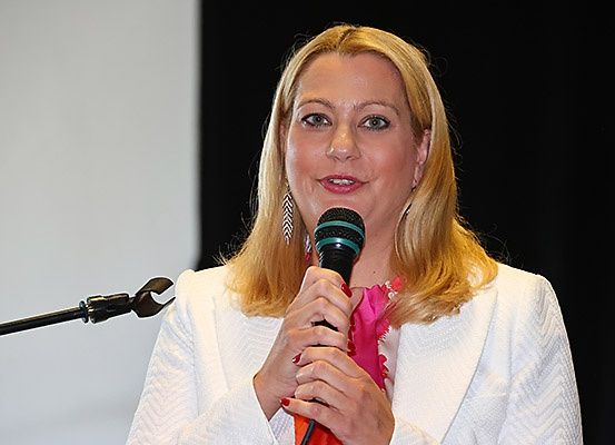 Eine Frau mit blonden Haaren spricht in ein Mikrofon, lächelt und hat einen schwarzen Gegenstand auf der linken Seite.