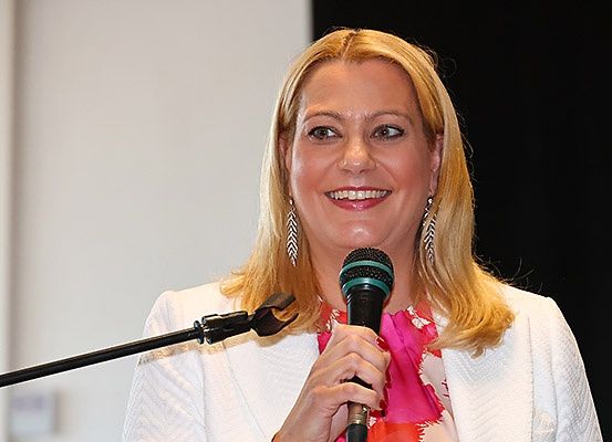 Eine Frau mit blonden Haaren spricht in ein Mikrofon und lächelt. Sie ist in einem weißen Blazer und mit Ohrringen gekleidet. Das Mikrofon wird in ihrer rechten Hand gehalten.
