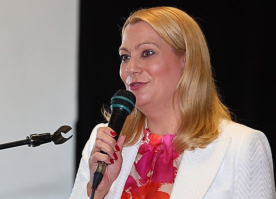 Eine Frau mit blonden Haaren spricht in ein Mikrofon und trägt einen weißen Blazer und einen rosa Schal.