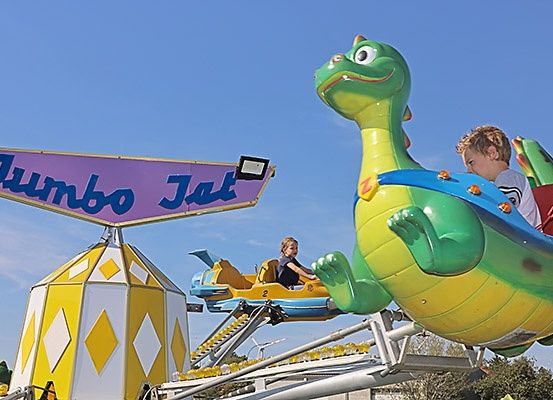 Zwei junge Kinder fahren eine bunte, dinosaurusförmige Attraktion in einem Vergnügungspark, die Jumbo Jet heißt.