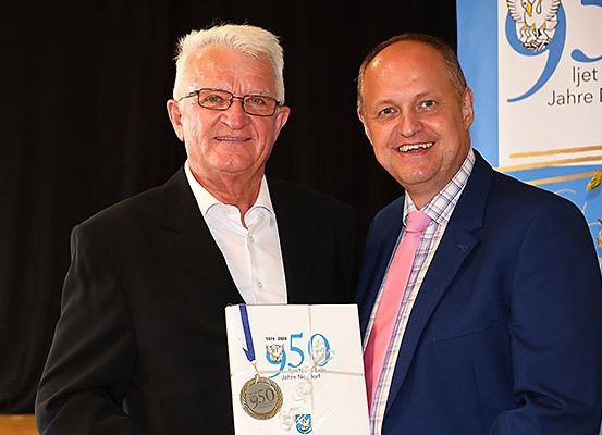 Zwei Männer in Anzügen stehen zusammen, einer hält ein Zertifikat mit der Nummer 950. Sie lächeln, was auf eine Preisverleihung hindeutet. Im Hintergrund befindet sich ein Banner mit Logo und Text.