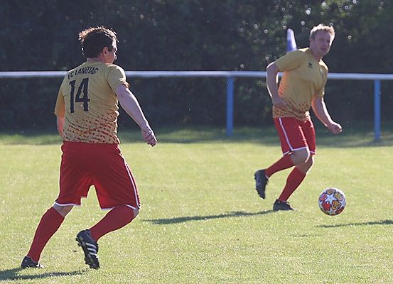 Zwei Fußballspieler in gelben und roten Trikots spielen auf einem Feld. Spieler 14 läuft auf den Ball zu.