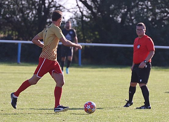 Ein Fußballspieler in einem gelben Trikot tritt einen Ball auf einem Feld, während ein Schiedsrichter in der Nähe zusieht.