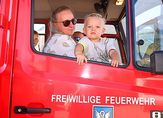 Eine Frau und ein Kind in einem roten Feuerwehrfahrzeug mit 'Freiwillige Feuerwehr' an der Seite. Die Frau trägt eine Sonnenbrille und das Kind lehnt sich aus dem Fenster.