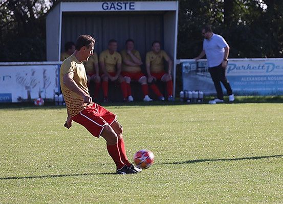 Ein Fußballspieler in roten Uniformen tritt einen Ball auf einem grünen Feld. Vier Personen sitzen auf einer Bank hinter ihm. Sie tragen gelbe Uniformen. Ein Mann steht neben einer Wand mit Werbetafeln.