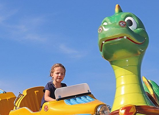 Ein kleines Mädchen fährt eine gelbe Auto-ähnliche Attraktion, wahrscheinlich in einem Vergnügungspark, vor einem klaren blauen Himmel.