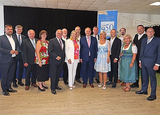 Eine Gruppe von Menschen in formeller Kleidung steht vor einem Banner, das '50 Jahre' feiert. Das Banner ist blau, und sie stehen auf einem Holzboden mit schwarzem Vorhang im Hintergrund.