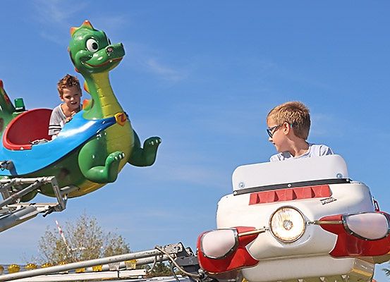 Zwei junge Jungen fahren auf einem Jahrmarkt eine farbenfrohe Drachen- und eine Raketenattraktion bei blauem Himmel.