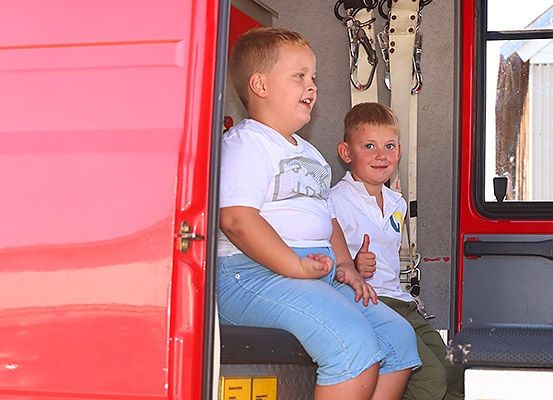Zwei junge Jungen sitzen in einem roten Feuerwehrauto, lächeln und zeigen Daumen hoch. Das Fahrzeug hat gelbe und rote Ausrüstung.