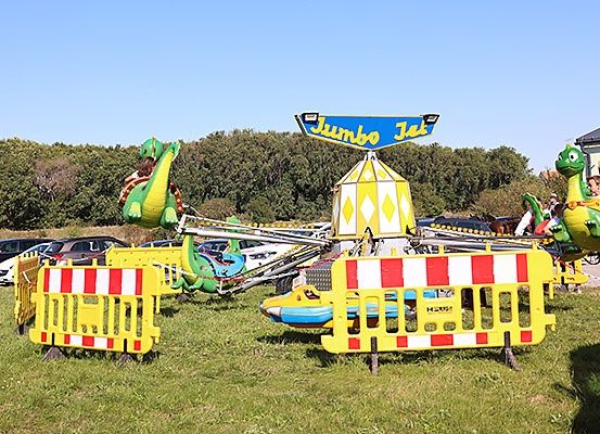 Ein Kinderfahrgeschäft namens Jumbo Jet mit Drachen-Motiven steht auf einem Rasen. Barrieren umgeben die Attraktion und ein paar Leute sind in der Nähe. Bäume und geparkte Autos sind im Hintergrund.
