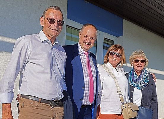 Vier Personen, zwei Männer und zwei Frauen, stehen zusammen draußen. Sie lächeln und scheinen für ein Foto zu posieren. Die Männer tragen Anzüge und Krawatten, während die Frauen lässig gekleidet sind. Sie stehen vor einem Gebäude mit blauer Wand.