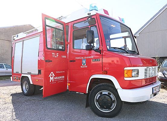 Ein roter Feuerwehrwagen mit der Aufschrift 'TLF 1000' und 'FREIWILLE FEUERWEHR NEUDORF' steht auf einem Schotterplatz. Dahinter ist ein Gebäude zu sehen, und eine Person steht daneben. Autos sind hinter dem Wagen geparkt.