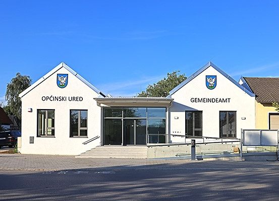 Ein weißes Gebäude mit Glastüren und Fenstern zeigt die Texte 'Opcinski Ured' und 'Gemeindeamt' über dem Eingang.