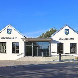 Ein weißes Gebäude mit Glastüren und Fenstern zeigt die Texte 'Opcinski Ured' und 'Gemeindeamt' über dem Eingang.
