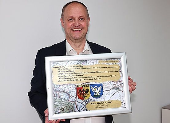 Ein Mann hält ein gerahmtes Zertifikat, lächelt und steht vor einer weißen Wand. Das Zertifikat hat eine Karte, Logos und Text.