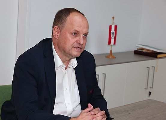 Ein Mann in einem dunklen Anzug sitzt an einem Schreibtisch, schaut in die Kamera, mit einer Flagge hinter ihm und Büchern auf einem Regal.