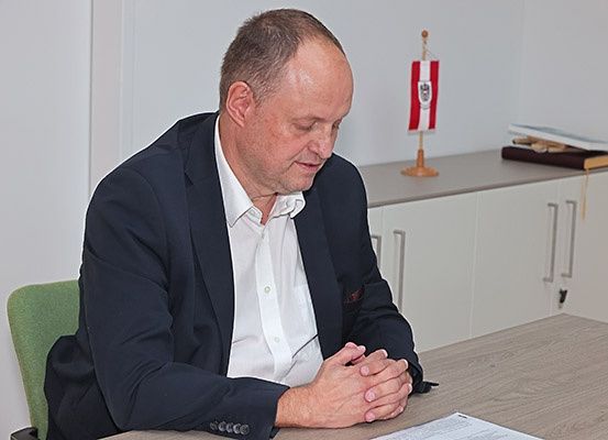 Ein Mann sitzt an einem Schreibtisch, nach vorne gerichtet, in einem dunklen Anzug. Er hat die Hände ineinander gefaltet. Hinter ihm befinden sich eine Flagge und ein Schrank.