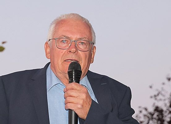 Ein älterer Mann mit Brille hält ein Mikrofon und scheint bei einer Veranstaltung im Freien zu sprechen.