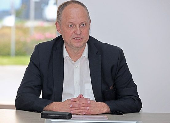 Ein Mann in einem Anzug sitzt mit verschränkten Händen an einem Schreibtisch, anscheinend in einem Konferenzraum.