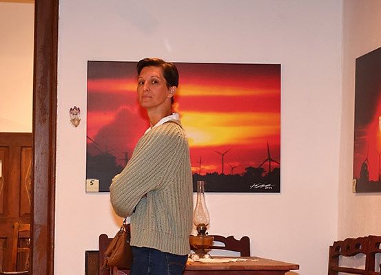 Eine Frau steht in einem Raum mit einem Sonnenuntergangsbild an der Wand, einer Lampe auf dem Tisch und Holzstühlen.