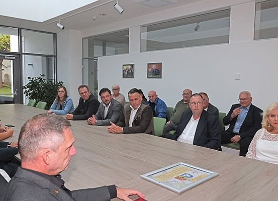 Eine Gruppe von Menschen sitzt an einem langen Tisch in einem Besprechungsraum mit Glaswänden. Einige schauen den Mann auf der linken Seite an, der auf etwas auf dem Tisch schaut.