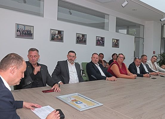 Eine Gruppe von Menschen, einige in Anzügen, sitzt um einen Tisch herum. Papiere, ein Telefon und ein gerahmtes Bild liegen auf dem Tisch. Der Raum hat eine Glaswand, Deckenlampen und gerahmte Fotos an der Wand.