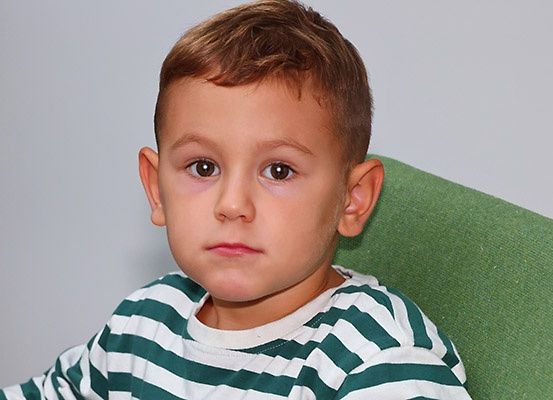 Ein junger Junge mit kurzen braunen Haaren und braunen Augen, der ein weiß-grün gestreiftes T-Shirt trägt, sitzt auf einem grünen Stuhl und schaut mit leicht geöffnetem Mund nach vorne.