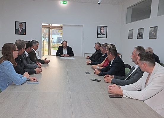 Eine Gruppe von Personen, sowohl Männer als auch Frauen, sitzen um einen langen Tisch herum, wahrscheinlich in einer Besprechung. Einige führen Gespräche.
