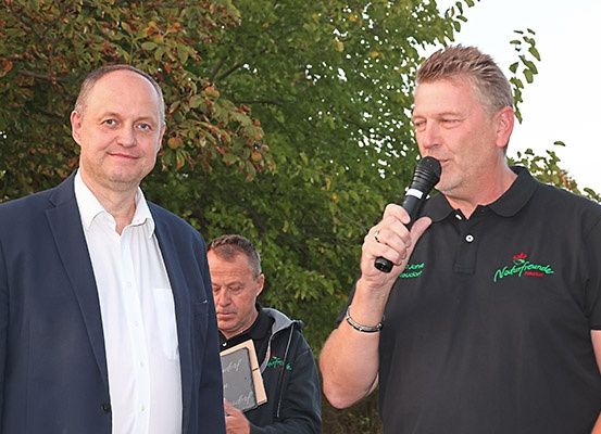 Drei Männer stehen draußen mit Bäumen im Hintergrund. Der Mann in der Mitte hält ein Mikrofon und spricht. Der Mann auf der linken Seite trägt einen Anzug und hält eine gerahmte Plakette. Der Mann auf der rechten Seite trägt ein schwarzes Polohemd mit grünem Text.