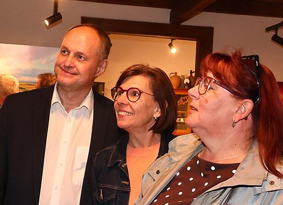 Drei Menschen stehen zusammen und lächeln für ein Foto. Der Mann trägt einen Anzug und die Frauen tragen eine Brille und Jacken. Ein Gemälde und eine Lampe sind im Hintergrund.