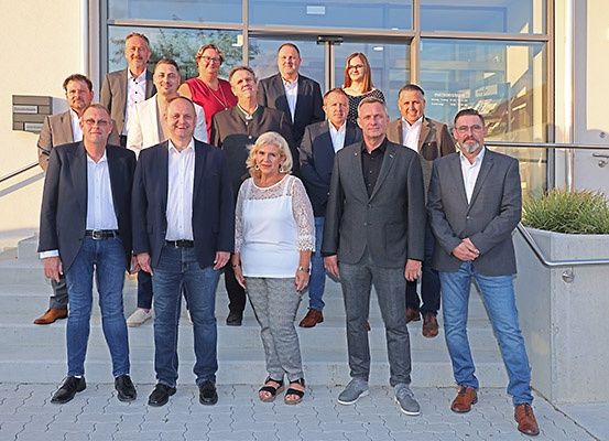 Eine Gruppe von Menschen in formeller Kleidung, die vor einem Gebäude mit Glaswänden und Treppen für ein Foto posiert.