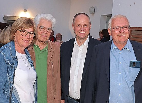 Vier Erwachsene, zwei Männer und zwei Frauen, lächeln und posieren für ein Foto. Sie tragen Brillen und formelle Kleidung. Hinter ihnen stehen weitere Menschen und ein Raum mit weißen Wänden.
