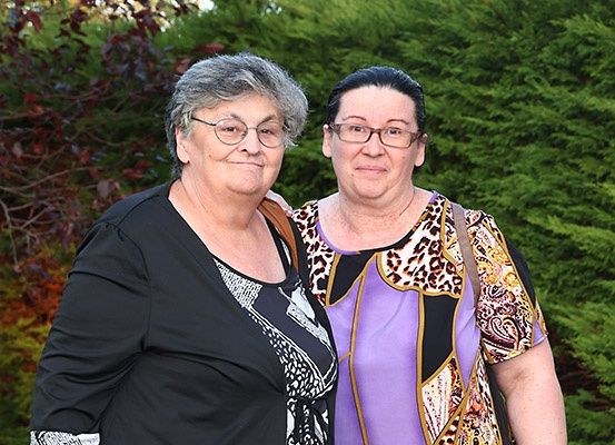 Zwei Frauen stehen Seite an Seite im Freien vor einem grünen Busch. Die Frau auf der linken Seite trägt eine Brille und ein schwarzes Oberteil. Die Frau auf der rechten Seite trägt ein Leopardenprint-Oberteil.