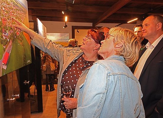 Mehrere Menschen versammeln sich in einem Raum und betrachten ein großes Gemälde an der Wand. Eine Frau zeigt auf das Gemälde, während eine andere Frau ein Weinglas hält. Der Mann auf der rechten Seite trägt einen Anzug und schaut zu.