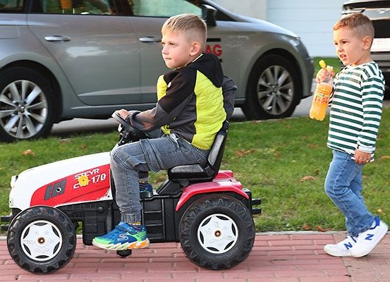 Zwei junge Jungen, einer fährt einen Spielzeugtraktor, der andere hält eine Flasche, sind draußen in der Nähe geparkter Autos.