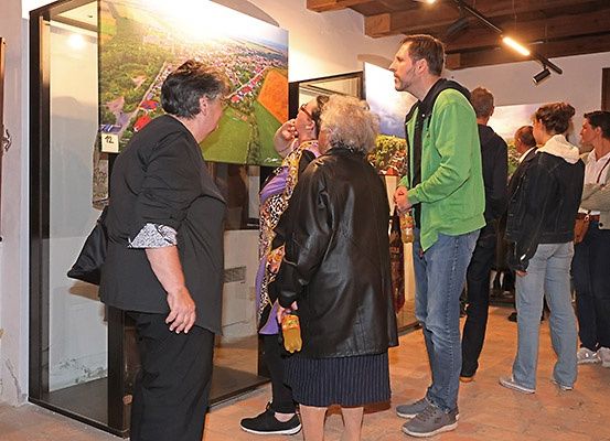 In einem Raum mit einem Gemälde in einer Glasvitrine stehen eine Gruppe von Menschen und unterhalten sich. Eine Frau hält eine Flasche. Der Mann im grünen Pullover schaut sie an.