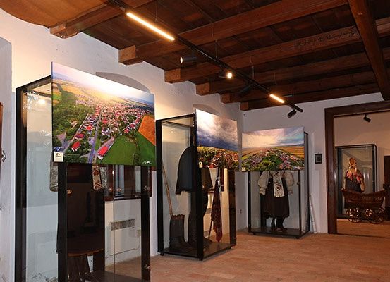 Ein Raum mit Holzbalken und gefliestem Boden, der Glasdisplays mit traditioneller Kleidung und Landschaftsfotografien aufweist.