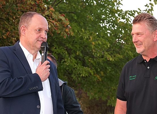 Zwei Männer stehen im Freien, einer mit einem Mikrofon, lächeln vor grünen Bäumen.