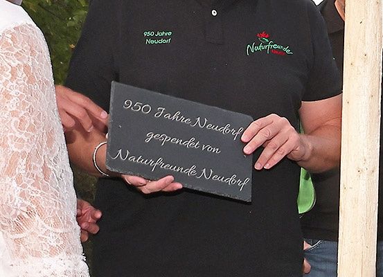 Eine Person hält eine Schiefertafel mit der Aufschrift '950 Jahre Neudorf' vor einer Gruppe von Menschen, möglicherweise während einer Feier.