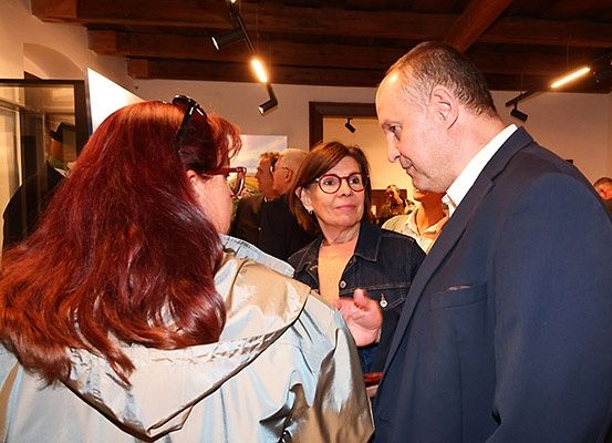 Ein Mann in einem Anzug spricht mit zwei Frauen in einer Galerie. Die Frauen tragen Brillen und Jacken. Scheinwerfer hängen von der Decke. Kunstrahmen sind im Hintergrund zu sehen.