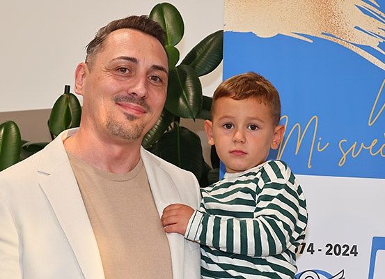 Ein Mann und ein junger Junge stehen lächelnd zusammen, mit einer Pflanze im Hintergrund. Dahinter zeigt ein blau-weißer Banner '714 - 2024' und einige goldene Texte.
