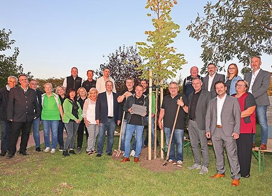 Eine Gruppe von Menschen steht in einem Park, einige pflanzen einen Baum. Sie sind in formeller Kleidung gekleidet, einige halten Schaufeln und andere tragen Brillen. Der Park hat Gras und Bäume, mit Bänken und einer Betonmauer im Hintergrund.
