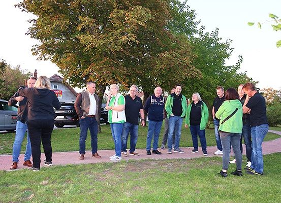 Eine Gruppe von Menschen in Grün und Schwarz steht in einem Park, unterhält sich und ist von Bäumen und geparkten Autos umgeben.
