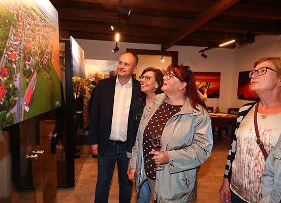 Eine Gruppe Erwachsener, darunter zwei Frauen und ein Mann, betrachten eine Luftaufnahme einer Stadt, die an einer Wand in einer Kunstgalerie ausgestellt ist.