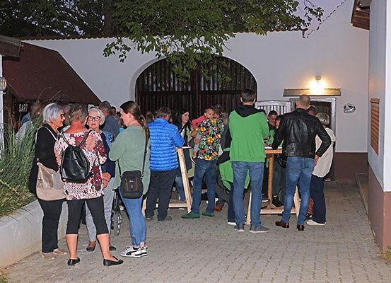 Eine Gruppe von Menschen steht vor einem Gebäude mit einem Bogen, möglicherweise in einer Schlange wartend. Bäume und Pflanzen sind in der Umgebung.